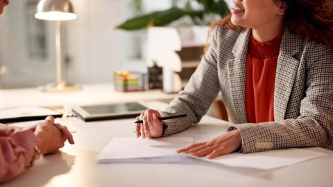 Two people sit across a desk from each other. One holds a pen and there are papers on the desk.