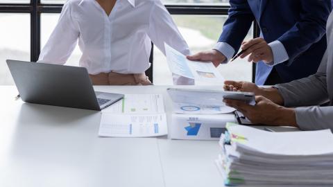 Three people gather around a table, looking at a laptop and pages printed with graphs and charts.