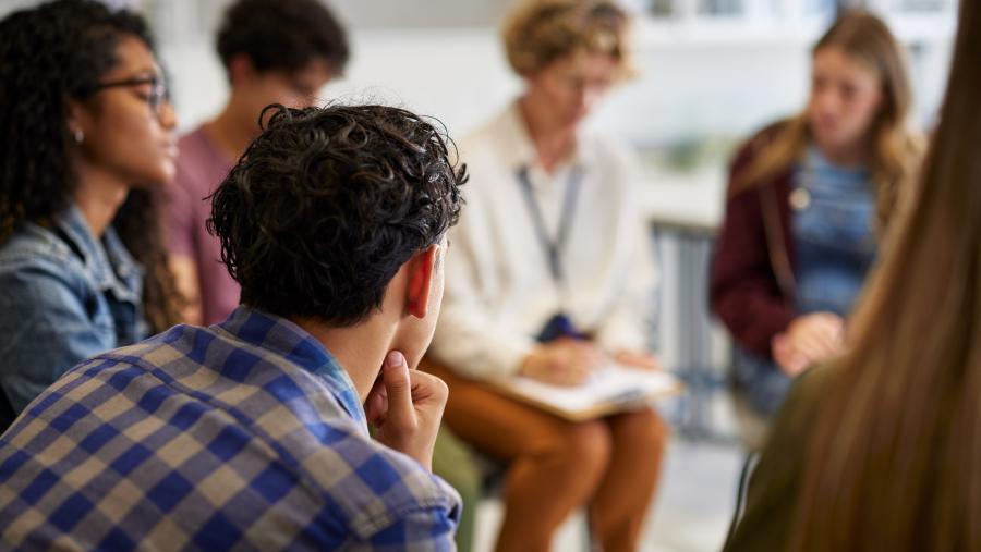 A group of young adults sit in a circle and one woman is writing on a clipboard.