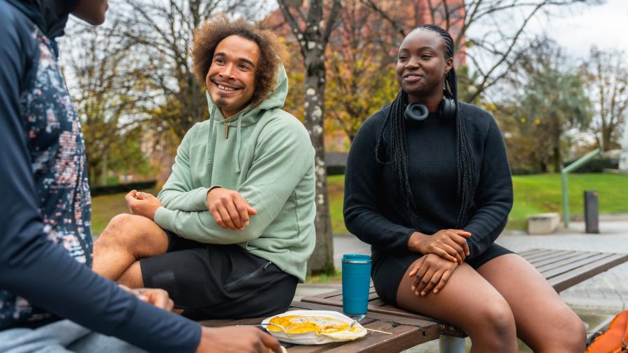 Three young adults sit on a bench, share food, and smile at each other.