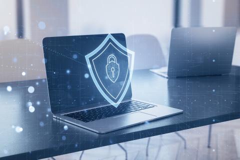 Laptop with a digital shield symbol onscreen signifies cybersecurity concept, blended with an office interior background.