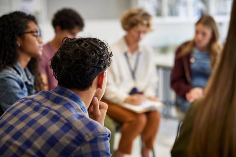 A group of young adults sit in a circle and one woman is writing on a clipboard.