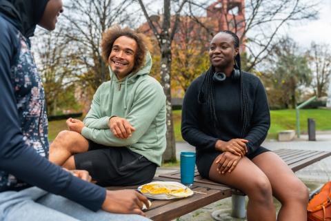 Three young adults sit on a bench, share food, and smile at each other.